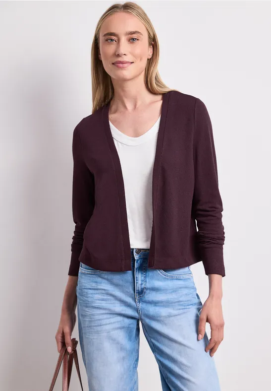 Vrouw draagt wit T-shirt, open bordeaux vest, lichte jeans en houdt een bruine handtas vast.