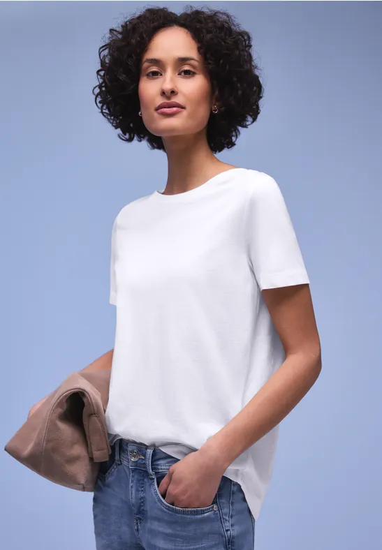 Persoon voor een neutrale achtergrond met wit t-shirt, spijkerbroek en houdt een jas in de hand.