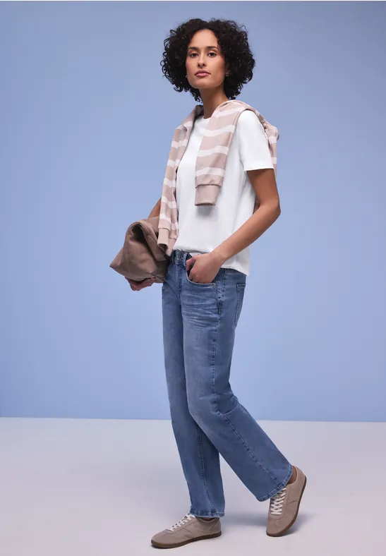 Persoon draagt T-shirt, spijkerbroek, sjaal, sneakers en handtas voor een lila achtergrond.