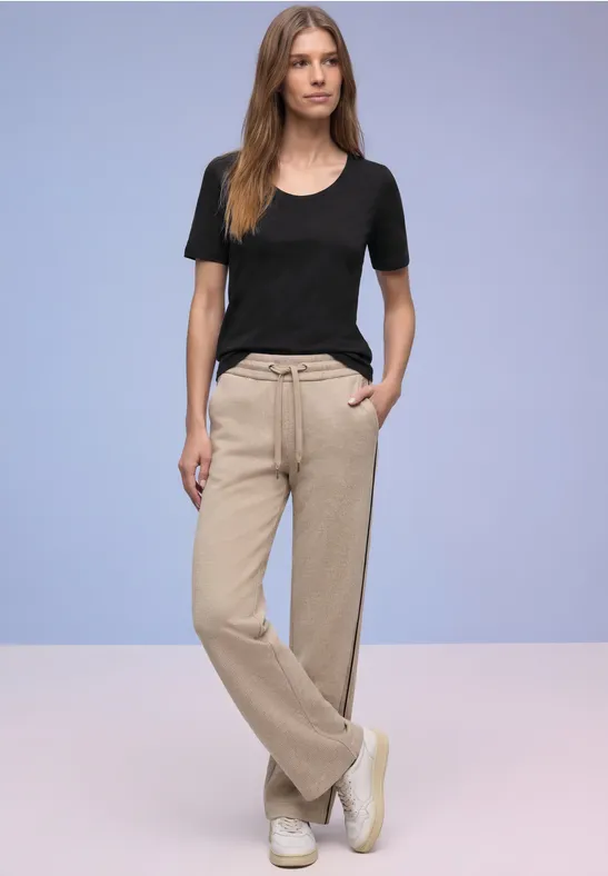 Frau trägt schwarzes T-Shirt, beige weite Hose und weiße Sneaker vor neutralem Hintergrund.