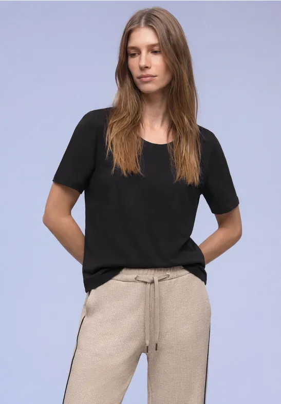 Frau mit langen Haaren trägt schwarzes T-Shirt und beige Hose vor hellviolettem Hintergrund.