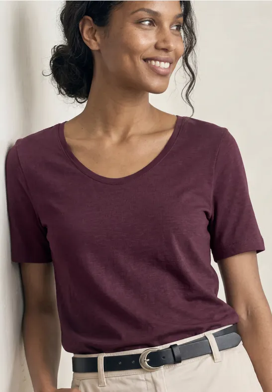 Een glimlachende vrouw in een bordeauxrood T-shirt met korte mouwen en een zwarte riem.