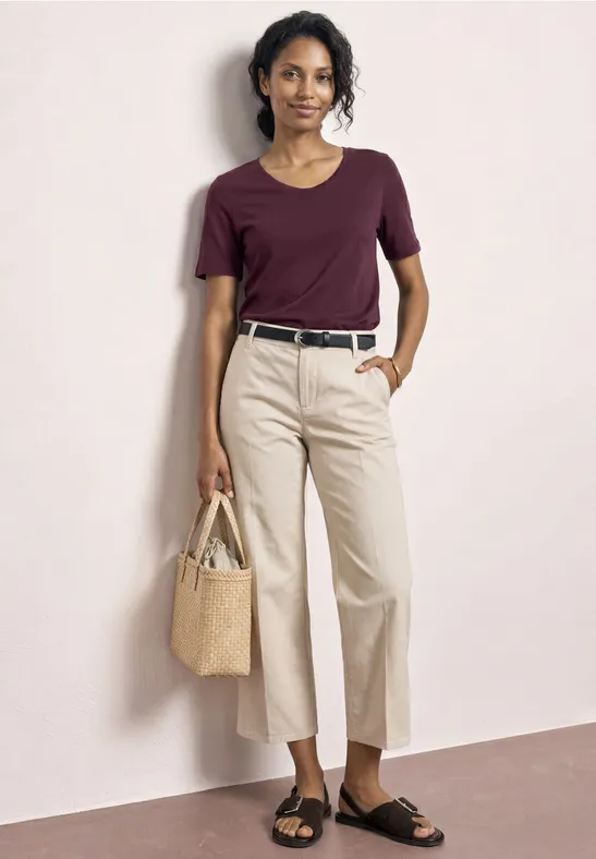 Een vrouw draagt een bordeaux t-shirt, lichte broek, zwarte riem, sandalen en een strooien tas.