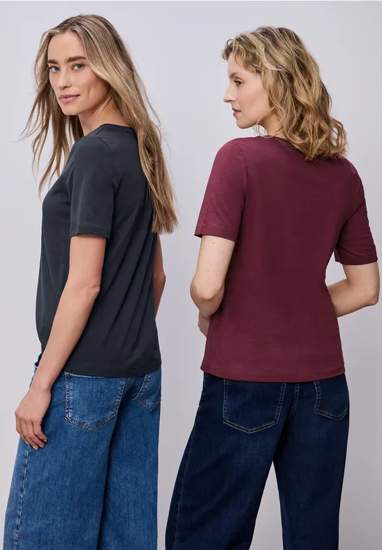 Zwei Frauen in legeren T-Shirts und Jeans stehen Rücken an Rücken, Blick seitwärts.