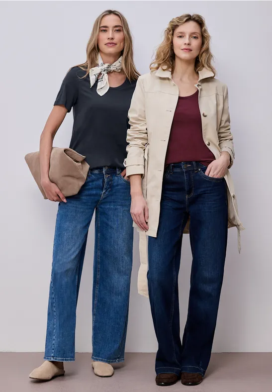 Zwei Frauen in Jeans, eine mit schwarzem Shirt und Halstuch, die andere mit beigem Trenchcoat, vor hellem Hintergrund.