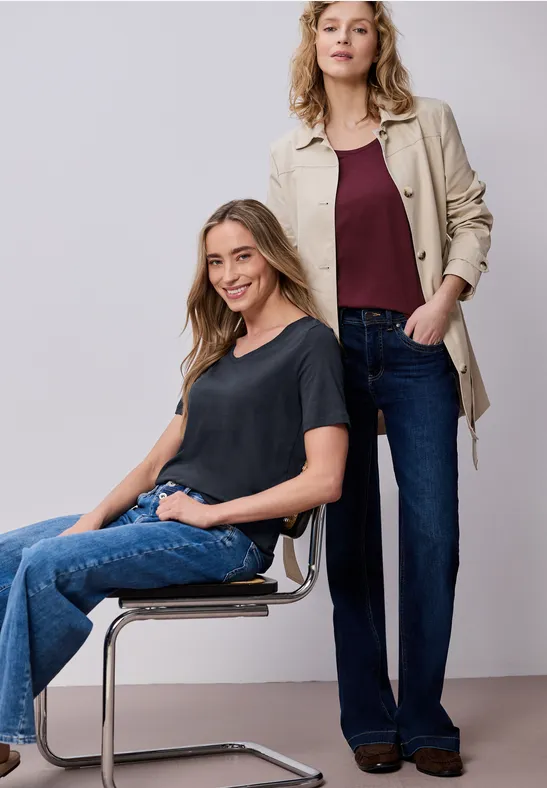 Zwei Frauen in Jeans und lässigen Oberteilen, eine sitzt auf einem Stuhl, die andere steht dahinter.
