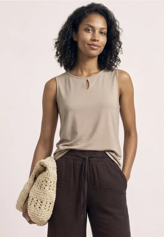 Vrouw in beige top en bruine broek draagt gehaakte tas voor een witte achtergrond.