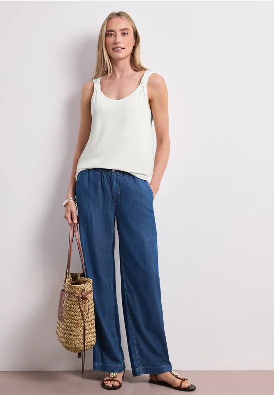 Frau in weißem Top, weiten blauen Hosen, schwarzen Sandalen und Korbtasche.