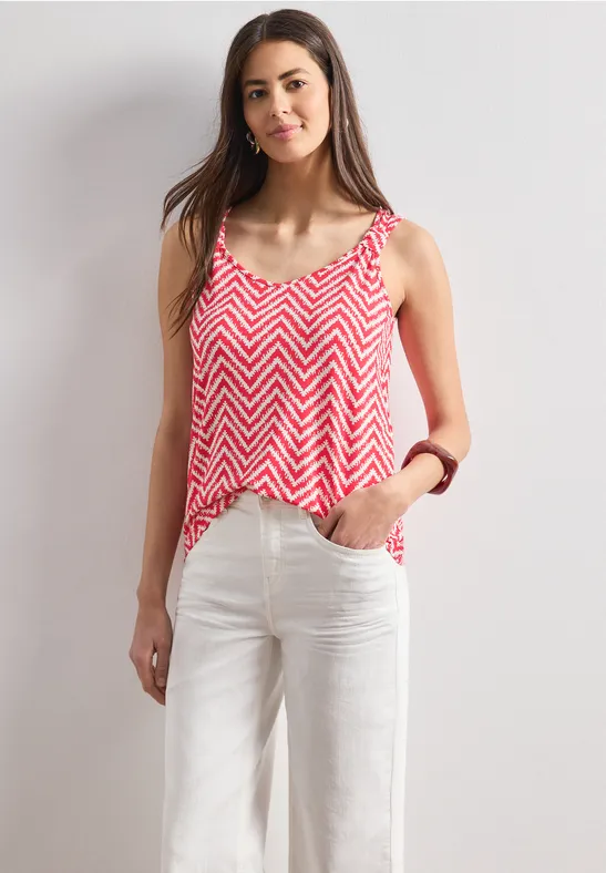 Vrouw in witte broek en rood-wit zigzagtop voor een neutrale achtergrond.
