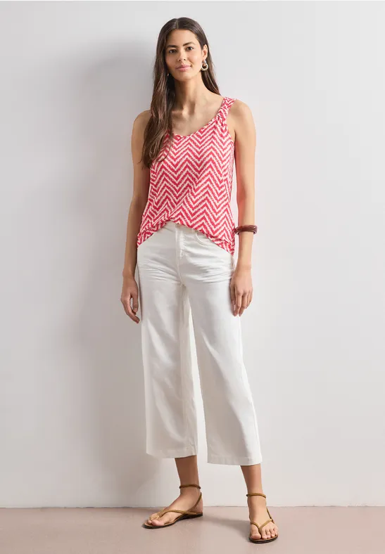 Vrouw in roze zigzagtop, witte brede cropped jeans en sandalen voor witte achtergrond.