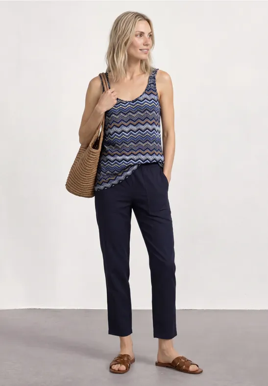 Vrouw met strooien tas, gemêleerd topje, donkerblauwe broek en bruine sandalen.