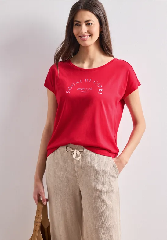 Een vrouw draagt een rood T-shirt en beige broek, houdt een tas vast voor een lichte achtergrond.