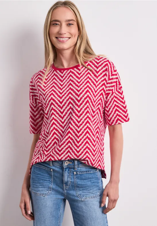 Vrouw in rood-wit zigzagshirt en blauwe jeans voor een lichte achtergrond.