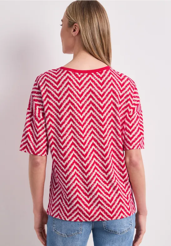 Vrouw van achter gezien in rood-wit zigzagshirt met driekwart mouwen en een blauwe spijkerbroek.