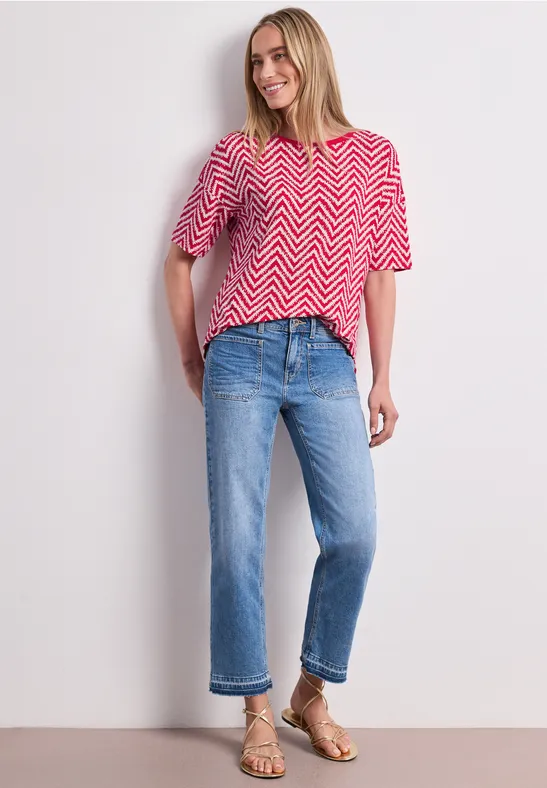 Vrouw met rood-wit zigzag T-shirt, blauwe cropped jeans en gouden sandalen voor een neutrale achtergrond.