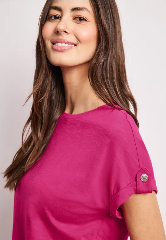 Frau mit langen Haaren in pinkem T-Shirt mit aufgekrempelten Ärmeln und Knopfdetail.