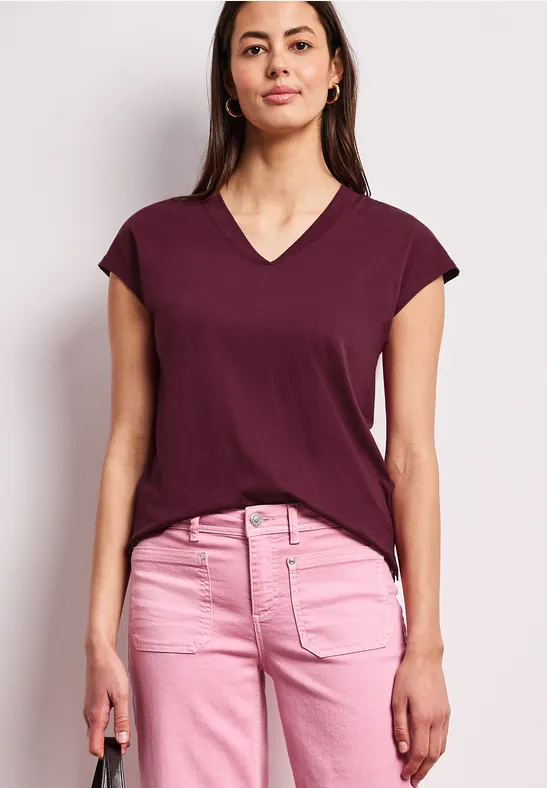 Frau trägt burgunderfarbenes T-Shirt mit V-Ausschnitt und rosa Hose, steht entspannt mit kleiner Handtasche.