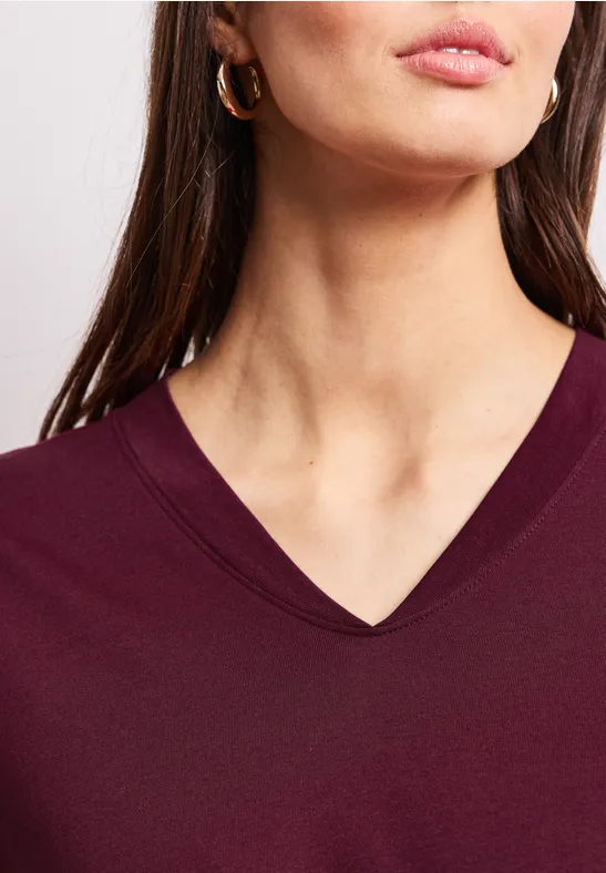 Frau trägt burgunderfarbenes V-Neck-Shirt und goldene Creolen, Oberkörper ab Brust sichtbar.