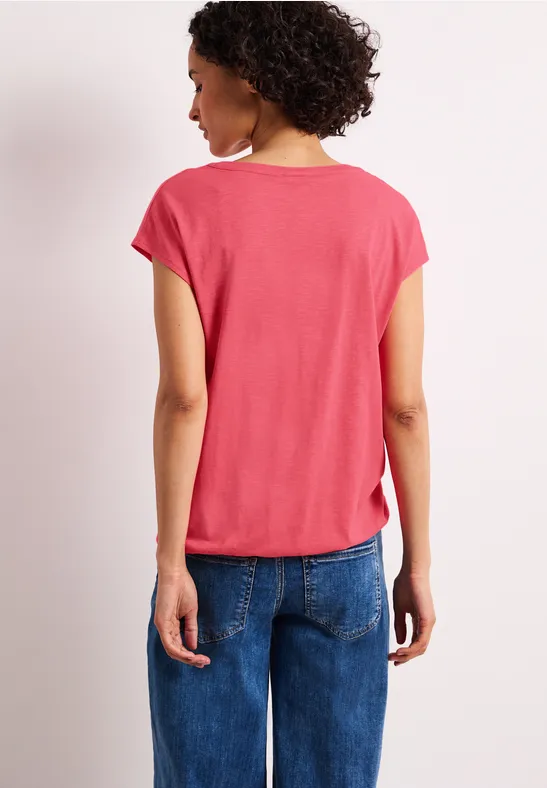 Frau von hinten in korallfarbenem T-Shirt und blauer Jeans, lockere Passform, Arme locker an den Seiten.
