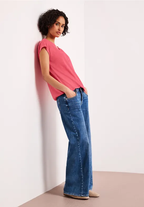 Frau lehnt an weißer Wand, trägt pinkes Oberteil, weite Jeans und bequeme Schuhe.