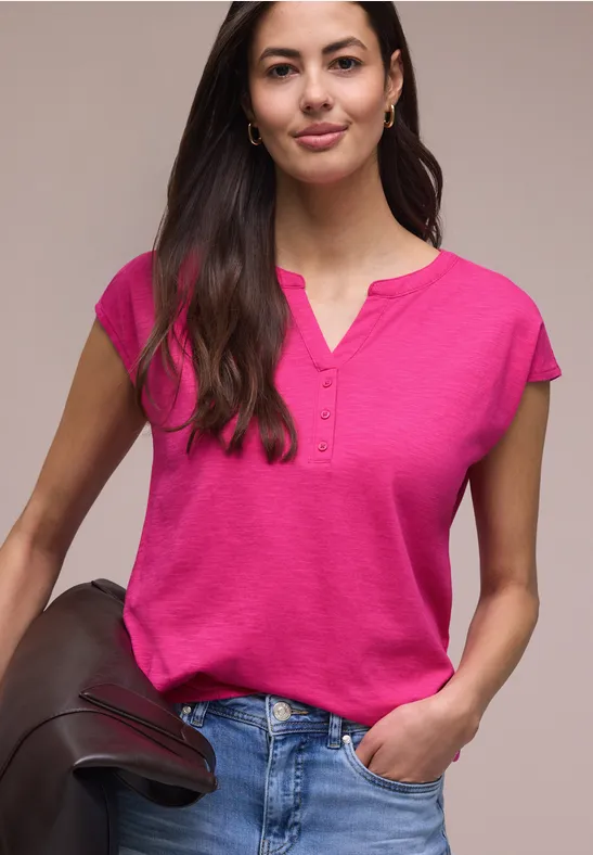 Frau in pinkem Henley-T-Shirt und Jeans, hält eine Lederjacke locker in der Hand.