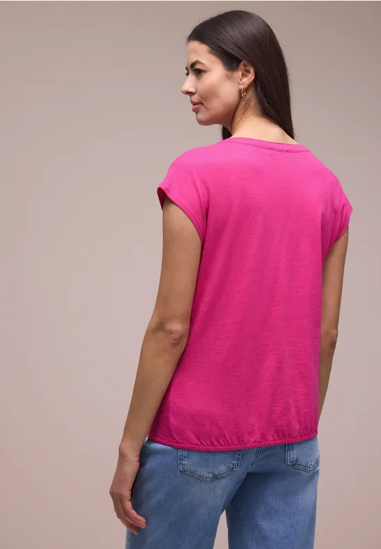Frau in pinkem T-Shirt und Jeans, von hinten aufgenommen, vor neutralem Hintergrund.