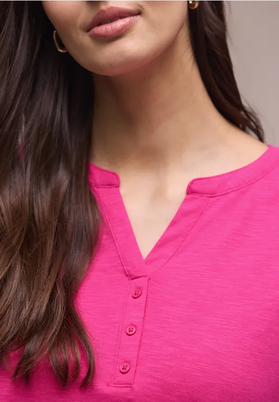 Frau mit langen dunklen Haaren trägt ein knallpinkes Shirt mit V-Ausschnitt und Knopfleiste.