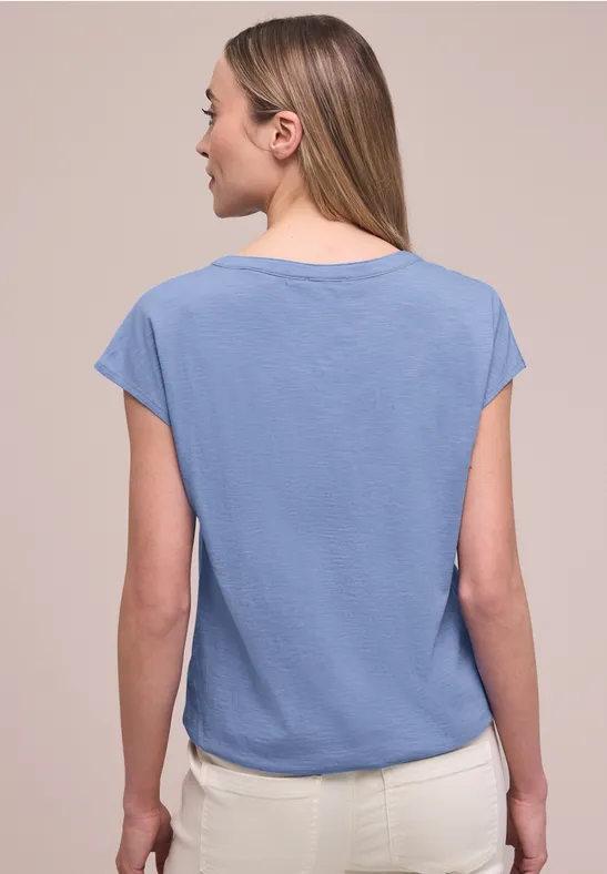 Eine Frau von hinten in hellblauem Kurzarmshirt und weißer Hose vor neutralem Hintergrund.