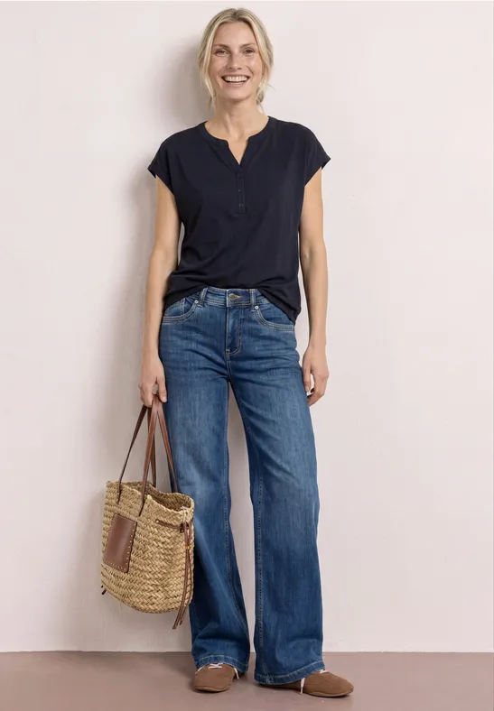 Frau in schwarzem T-Shirt und weiten Jeans hält eine Basttasche, steht vor hellbeigem Hintergrund.