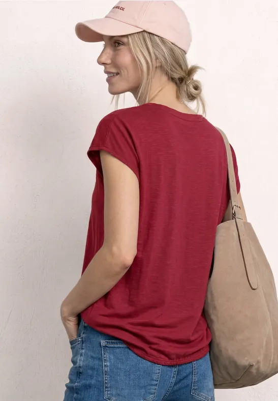 Frau von hinten in rotem T-Shirt, Jeans, rosafarbener Cap und beiger Tasche.