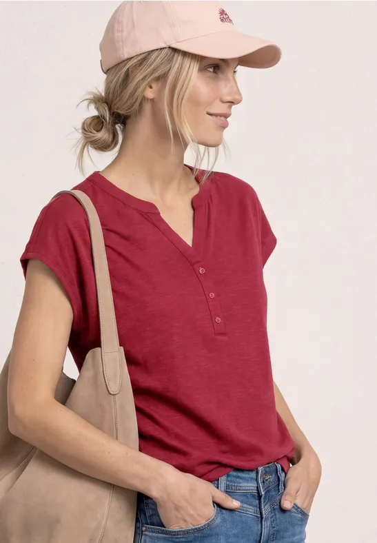 Frau mit rotem Kurzarmhemd, rosa Cap und beiger Schultertasche vor hellem Hintergrund.