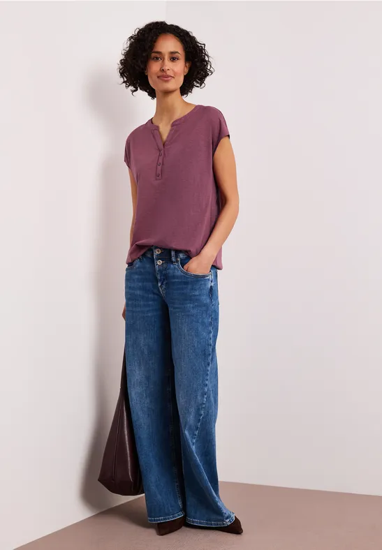 Een persoon staat voor een muur, met een paarse top en wijdvallende blauwe jeans.