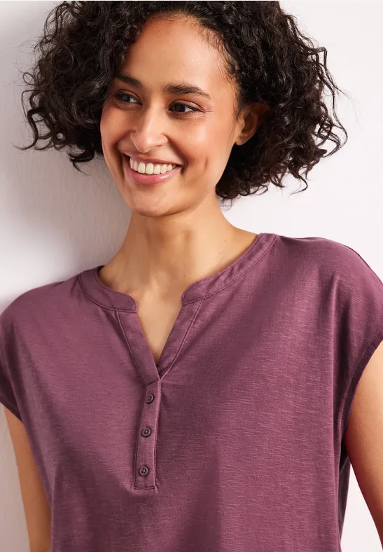 Persoon met krullend haar draagt een paars shirt met knopen voor een witte muur.