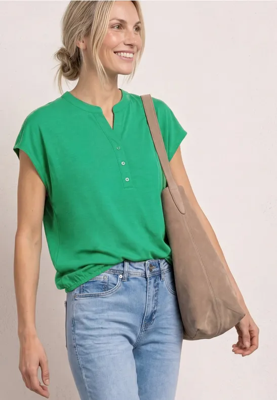 Frau in grünem Henley-T-Shirt, blauen Jeans und beiger Tasche vor neutralem Hintergrund.