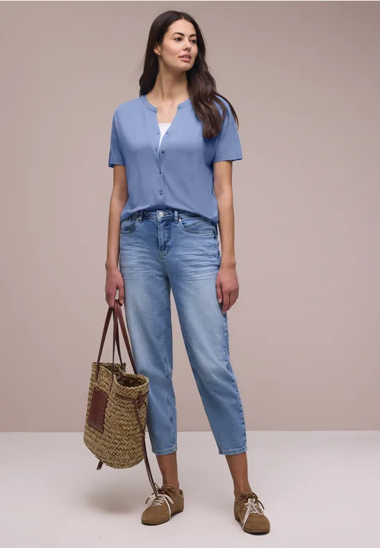 Frau in blauem Kurzarmshirt, weißem Top, blauen Jeans, mit Strohtasche und Loafers.