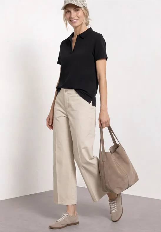 Eine Person steht in beigen Wide-Leg-Hosen, schwarzem Polo, trägt beige Sneaker und eine große Stofftasche.