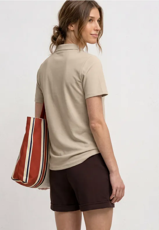 Eine Frau steht im Profil, trägt ein beiges Poloshirt, braune Shorts und eine rot-weiß gestreifte Tasche.