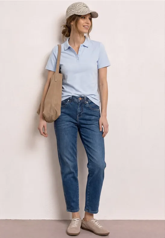 Frau steht vor hellem Hintergrund, trägt hellblaues Poloshirt, blaue Jeans, Sneakers und große Beuteltasche.