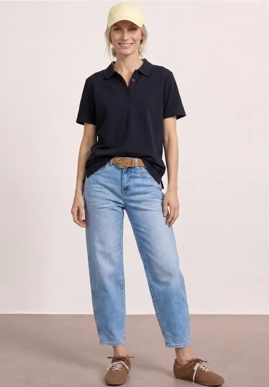 Femme en polo noir, jean clair taille haute, ceinture brune et chaussures en daim marron clair.