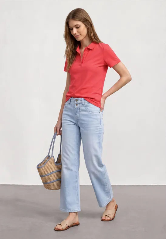Vrouw in rood poloshirt, lichtblauwe jeans en sandalen, met een gevlochten tas.