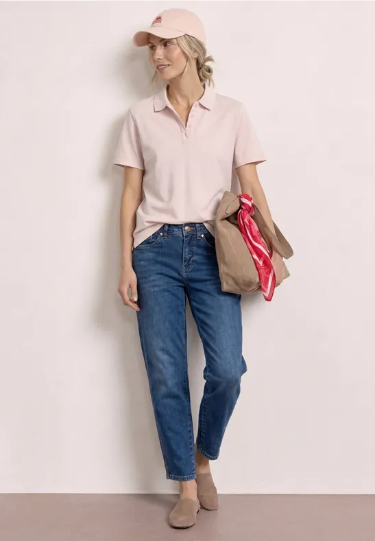 Femme debout devant un mur clair, en polo clair, jeans bleus et chaussures basses, tenant un sac rouge à la main.