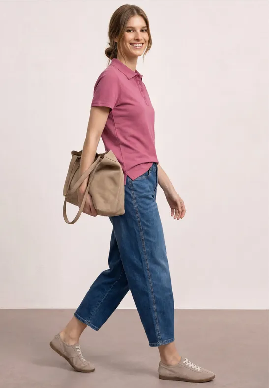 Eine Frau im Profil trägt ein rosafarbenes Poloshirt, blaue Jeans und eine Tasche über der Schulter.