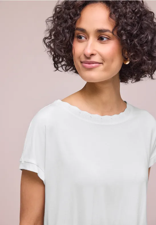 Person mit lockigem Haar trägt ein weißes T-Shirt mit wellenförmigem Halsausschnitt und umgekrempelten Ärmeln.