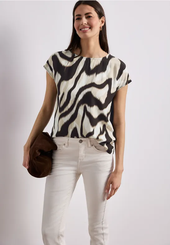 Frau in Zebratop und weißen Jeans mit braunem Hut vor neutralem Hintergrund.