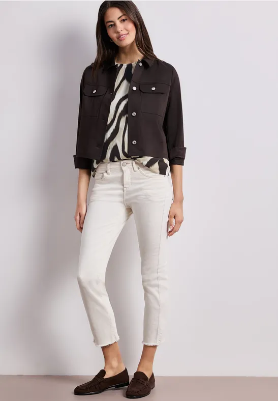 Dame in weißer Jeans, schwarzer Kurzjacke, gemusterter Bluse und braunen Loafers vor hellem Hintergrund.