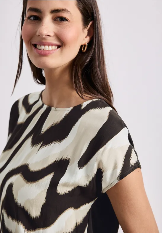 Person trägt kurzärmelige Bluse mit abstraktem Schwarz-Creme-Muster vor schlichtem Hintergrund.