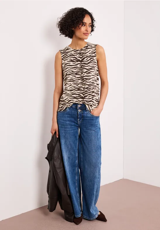 Vrouw in mouwloze zebraprint top, wijde jeans en zwarte jas voor een witte muur.