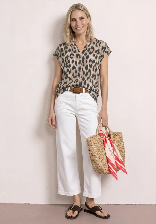 Vrouw in panterprint top, witte wijdere broek, zwarte sandalen en een strooien tas met rood sjaaltje.