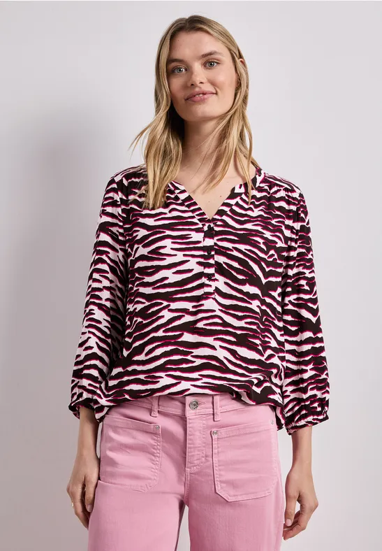 Eine Frau steht im rosa Outfit: Zebramuster-Bluse und rosa Hose vor hellem Hintergrund.