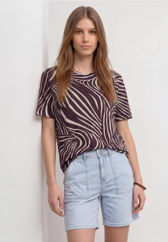 Persoon draagt een zebrapatroon T-shirt, lichte spijkershorts en een armband, staand voor witte achtergrond.